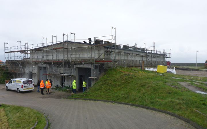 Ein Gebäude im Rohbau mit Gerüst. Davor stehen Menschen mit Warnjacken.