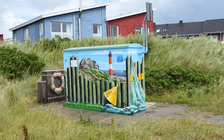 Trafostation bemalt mit dem Blick über den Zaun aufs Meer und den Leuchtturm